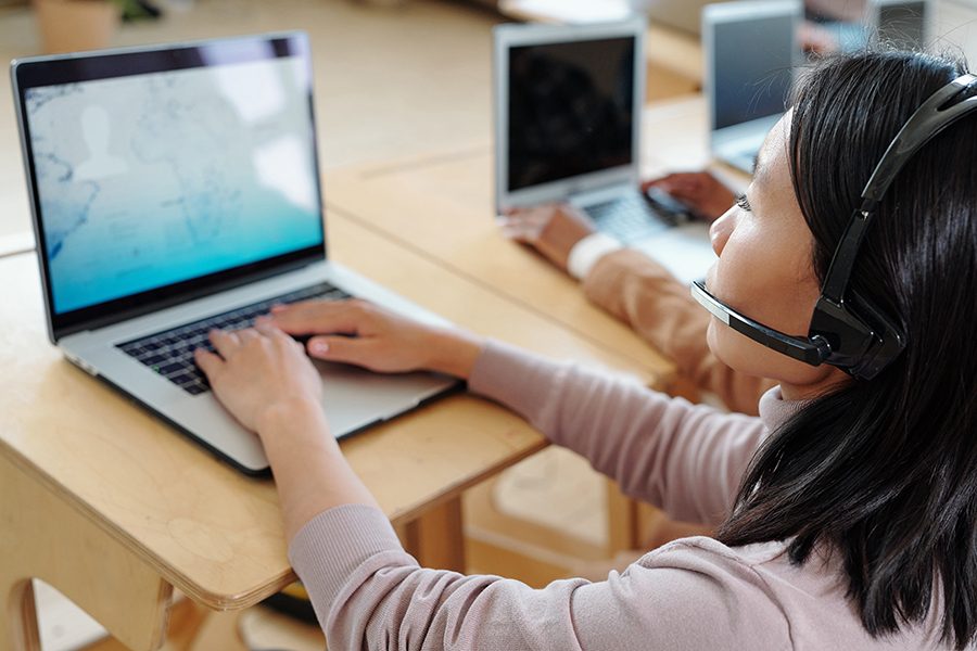 Over shoulder view of busy young Asian call center operator in wireless headset with microphone providing customer with information using laptop