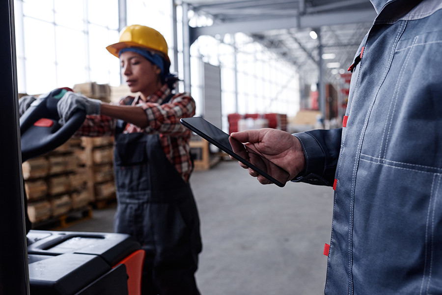 Hand of manager of quality control holding tablet against woman pushing cart Hand of male manager of quality control in workwear holding tablet against young woman pushing cart with packages