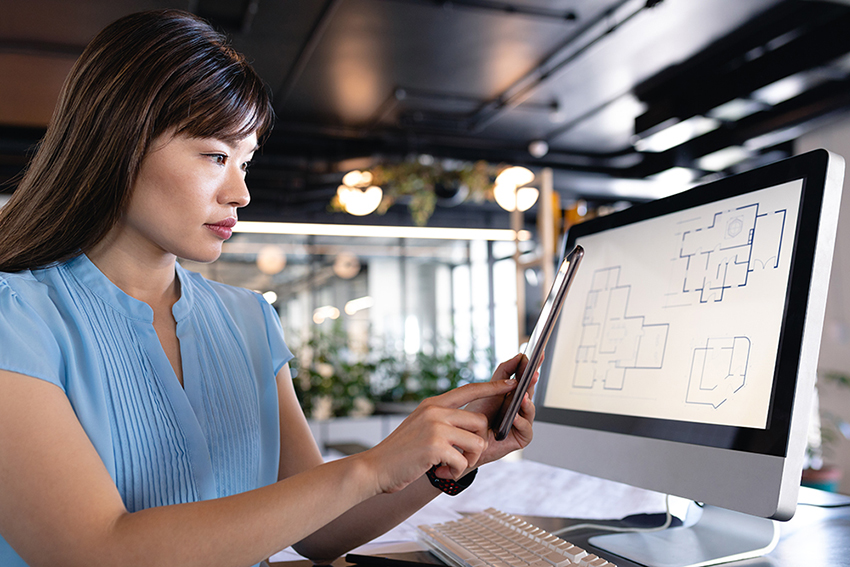 Side view of an Asian businesswoman wearing smart clothes and glasses, working in the modern office, sitting on a desk and using her smartphone and computer.